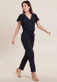 Tutina navy con scollo a V, maniche corte a volant, vita con laccio e gambe affusolate, abbinata a sandali con tacco alto dorati. Texture liscia.