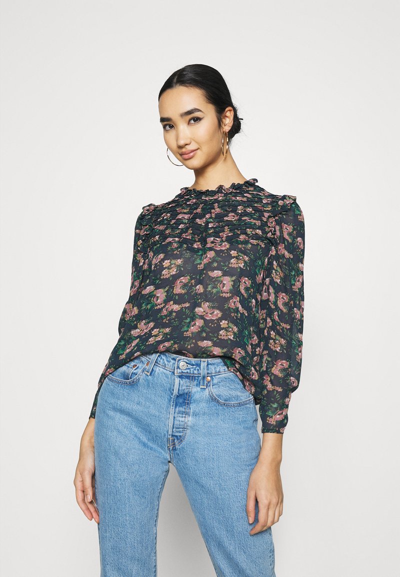 Blusa floreale scura con scollatura a volant e maniche lunghe, caratterizzata da motivi rosa e verdi, abbinata a jeans a vita alta di colore azzurro chiaro.