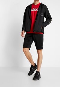 Svart lättviktsjacka med dragkedja och huva, röd t-shirt med vitt logo, svarta träningsshorts och svarta sneakers med strukturerad sula.