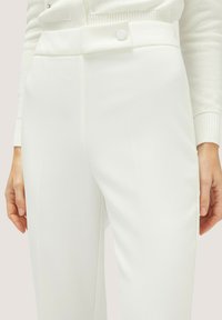 Pantaloni bianchi su misura con una texture liscia, dotati di chiusura laterale a bottone e dettaglio della piega frontale. Vita chiusa con una finitura pulita.