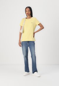 Tricou galben din bumbac, cu mâneci scurte, decolteu rotund și un mic logo brodat pe piept. Asortat cu blugi evazați albaștri și adidași albi.