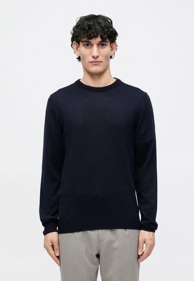 Pull en tricot bleu marine à manches longues, col rond et ourlet côtelé. Texture lisse sans motifs ni accents. Porté avec un pantalon gris clair.
