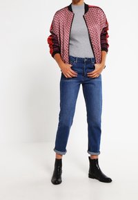 Red patterned bomber jacket with black accents, gray sweater, blue jeans rolled at cuffs, and black ankle boots.
