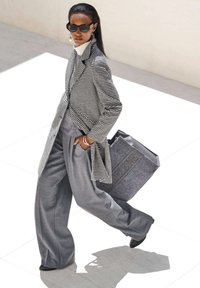 Een model draagt een zwart-wit hondentand blazer over een grijze outfit, terwijl ze een grijze vilten tas met reliëftekst vasthoudt. Slanke zonnebril maakt de look compleet.