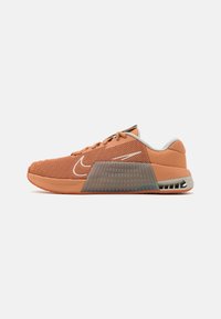 Pomarańczowy siateczkowy but sportowy z białym logo w kształcie łuku, grubą przezroczystą wkładką środkową oraz szarymi akcentami na pięcie i wewnętrznej wyściółce.