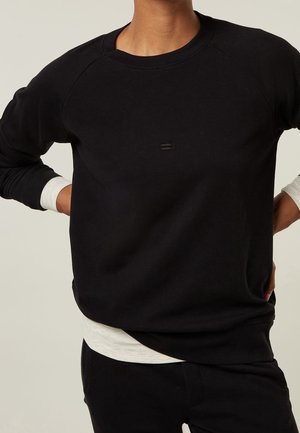 Personne portant un sweat-shirt noir à manches longues sur une chemise blanche, les mains posées sur les hanches, devant un fond uni.