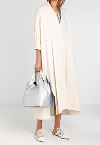 Lang, lichtbeige overhemdjurk met knopen, opgerolde mouwen en zijsplitten. Accessoires omvatten een zilveren handtas en witte instappers met perforaties.