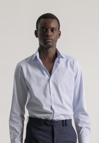 Camisa de vestir de rayas azul claro con una textura suave, mangas largas y cuello puntiagudo, combinada con pantalones oscuros ajustados.