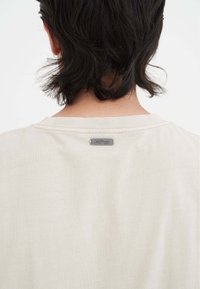 Hellbeige T-Shirt mit Rundhalsausschnitt, versehen mit einem kleinen metallischen Etikett am Rücken. Weicher, glatter Stoff mit einer schlichten Textur.