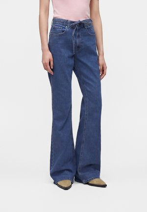 Personne portant un jean large taille haute bleu foncé avec une ceinture en tissu nouée à la taille et des chaussures pointues couleur beige.