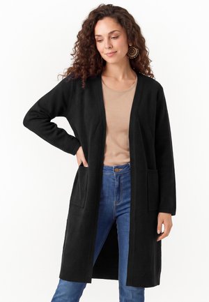 Frau mit lockigem Haar, die große runde Ohrringe, eine schwarze lange offene Strickjacke, ein beiges Top und blaue Jeans trägt, steht mit einer Hand in der Tasche.