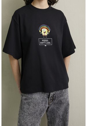 Person trägt schwarzes T-Shirt mit Cartoon-Figur, die einen Regenbogen hält, Text "WEIRD. DON'T CARE." und graue Acid-Wash-Jeans.
