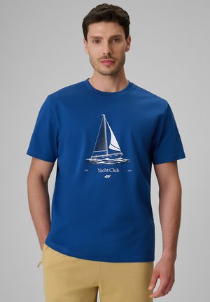 Mężczyzna ubrany w niebieski t-shirt z białym żaglówką i logo Yacht Clubu, w połączeniu z jasnobeżowymi spodniami, stoi na tle jednolitego szarego tła.