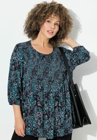 Blusa estampada con escote barco, detalles fruncidos y mangas de 3/4 en tonos verde azulado, negro y púrpura; combinada con un bolso tote negro.