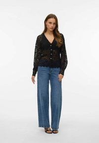 Cardigan in pizzo nero con scollo a V, maniche lunghe e chiusura con bottoni. Abbinato a jeans a gamba larga azzurri e sandali neri.