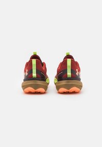 Een paar rood en oranje Under Armour hardloopschoenen met neon groene trekhaken, bruine tussenzolen en gestructureerde buitenzolen, getoond vanaf de achterkant.