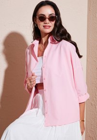 Light pink button-up shirt with roll-up sleeves, layered over a white top, worn with a white skirt. Smooth fabric, casual fit.