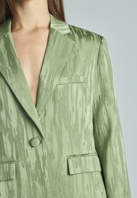 Blazer verde chiaro con un motivo testurizzato a onde. Chiusura con un singolo bottone e due tasche frontali. Finitura liscia simile alla satinata.