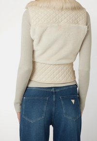 Veste matelassée beige avec col en fausse fourrure, portée sur un haut côtelé à manches longues, associée à un jean bleu taille haute avec des accents de poche.