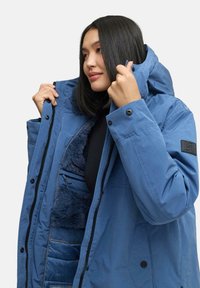 Veste isolante bleue avec une fermeture éclair à l'avant, un intérieur en fourrure et une capuche ajustable ; elle présente des boutons-pression et un logo sur la manche.