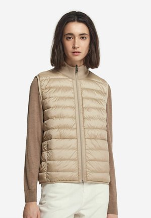 Beige Wattierte Weste mit hohem Kragen und Reißverschluss. Strukturierten Oberfläche mit gestepptem Muster, über einem braunen Langarmoberteil getragen.