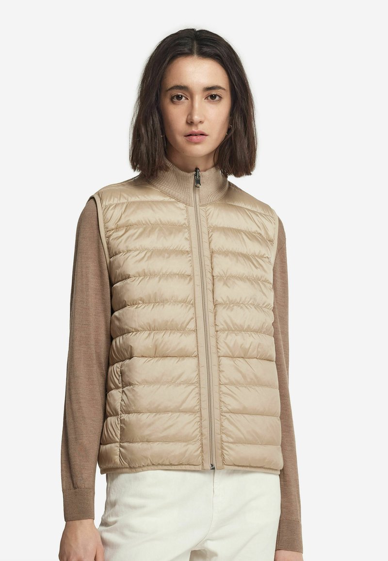 Vadderad väst i beige med hög krage och dragkedja. Texturerad yta med quiltat mönster, lager över en brun långärmad topp.