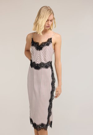 Blondynka w sukience midi bez rękawów, białej z pionowymi czerwonymi paskami i czarną koronkową lamówką, patrząca w dół, z jedną ręką zgiętą za plecami.