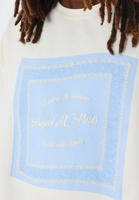 Sudadera crema con un gráfico azul claro que presenta patrones de paisley y texto bordado en blanco: "Explora lo desconocido, Project X Paris."