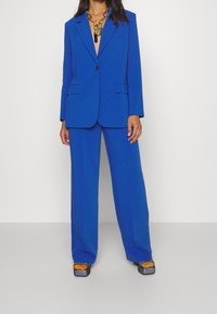 Kvinna iklädd kobaltblå blazer och vida byxor med kraftiga metalliska plattformsskor och lager av guldhalsband.