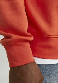 Oranje sweatshirtmouw met ribgebreide manchet; zachte textuur. Gelaagd over een wit shirt, gecombineerd met blauwe denim. Close-up focus op de stofdetails.