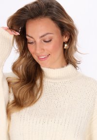 Pull en crème tricoté avec un col montant, mettant en valeur des accents en fil métallique, un tissu texturé et des manches volumineuses.