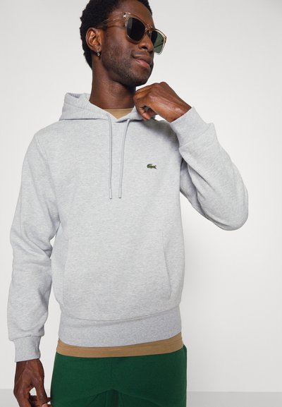 Hoodie gris avec cordons, poche avant et logo brodé vert. Porté sur une chemise beige, associé à un pantalon vert et des lunettes de soleil.