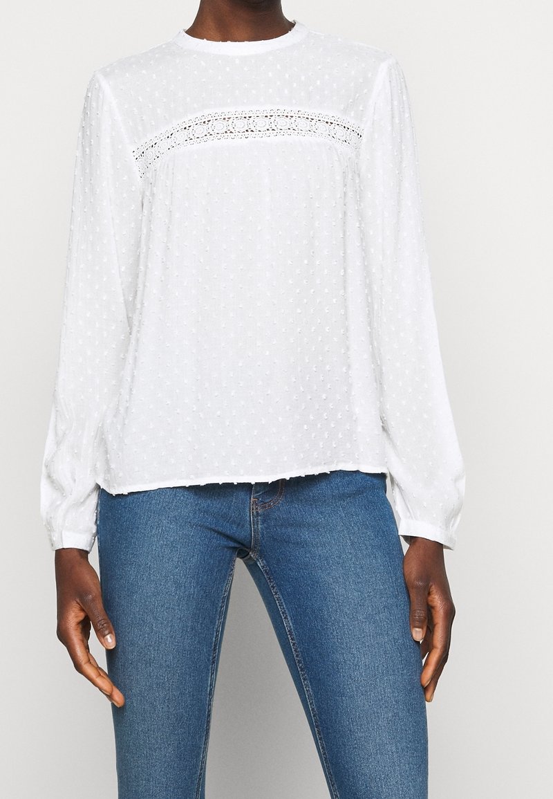 Blouse blanche à manches longues avec texture à pois et détail en dentelle sur la poitrine, portée avec un jean bleu.
