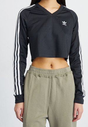 Femme portant un haut court Adidas noir à manches longues avec des bandes blanches sur les manches et un pantalon beige à taille élastique, debout devant un fond uni.