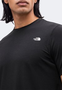 Άτομο που φοράει μαύρο T-shirt με στρογγυλή λαιμόκοψη και το λογότυπο The North Face στο αριστερό μέρος του στήθους, εμφανίζοντας την περιοχή του λαιμού και των ώμων.
