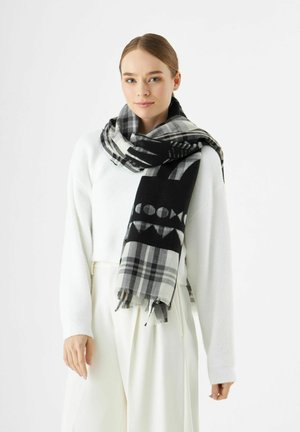 Jonge vrouw met lichtbruin haar draagt een zwart-wit gestreepte sjaal over een witte trui en een witte broek met hoge taille.