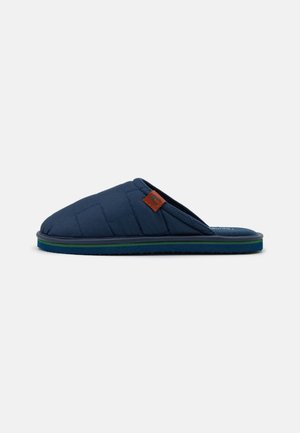 Mørkeblå quiltet slip-on tøffel med åben hæl, grønstribet sål og lille brun mærkat på siden, vist fra ydersiden.