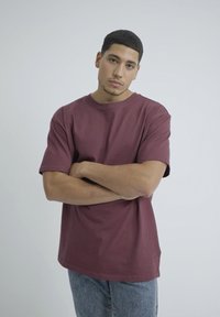 Burgundy pamut póló rövid ujjakkal, kerek nyakkivágással, laza szabással. A világoskék farmernadrágot viselő modell karba tett kézzel áll.