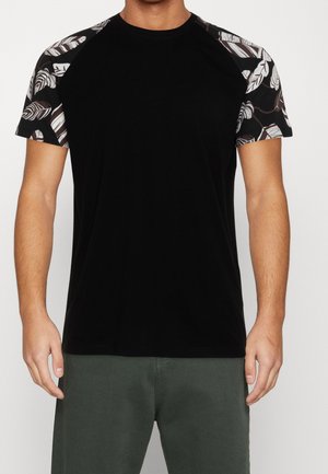 Homme portant un t-shirt noir à manches courtes avec des motifs de feuilles blanches et brunes sur les manches et un pantalon vert foncé, debout devant un fond uni.