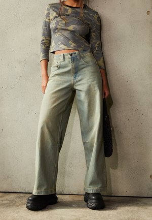 Osoba ubrana w zielony, kamuflażowy krótki top z długim rękawem, luźne, jasnoniebieskie jeansy z wysokim stanem, czarne masywne buty, stojąca na tle teksturowanej ściany.