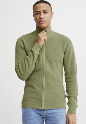Suéter verde de canalé con cierre de cremallera, cuello alto y mangas largas, que presenta un patrón de punto texturizado y un diseño ajustado.