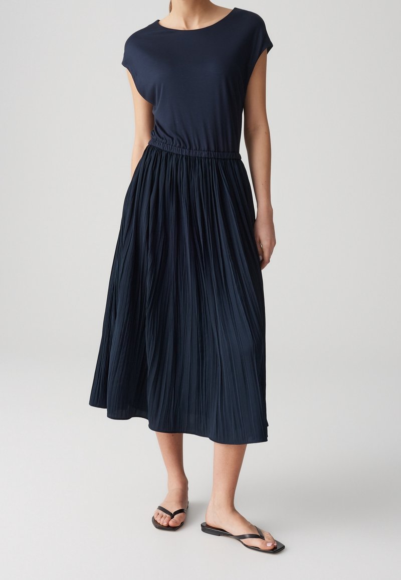 Femme portant un haut bleu marine à manches courtes avec une jupe midi plissée taille haute et des sandales plates noires sur un fond clair.
