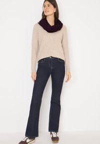 Pull beige à col large, associé à un jean évasé bleu foncé avec des coutures contrastées. Le modèle porte une écharpe en forme d'infini bordeaux et des baskets.