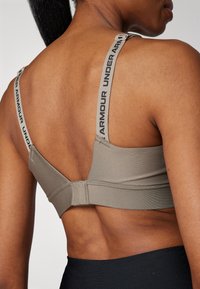 Grå sports-bh med justerbare stropper som har "UNDER ARMOUR"-tekst. Glatt stoff, enkelt design og sikker lukking bak.