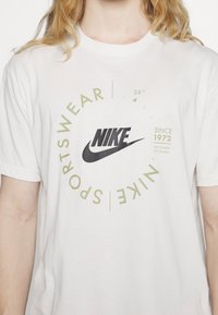 T-shirt en coton blanc avec logo Nike noir et design de texte circulaire vert avec les mentions "Sportswear" et "Depuis 1972". Manches courtes.