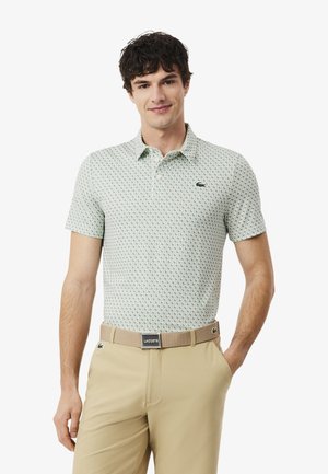 Joven con una camisa polo de color verde claro con estampado y pantalones beige con un cinturón beige de Lacoste, de pie con una mano en el bolsillo.