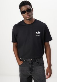 Crna pamučna majica s okruglim izrezom i bijelim Adidas logotipom na prsima, u kombinaciji s tamnim trapericama i crnim sunčanim naočalama.