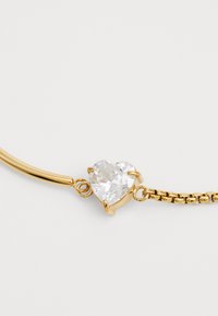 Χρυσό βραχιόλι με κεντρικό στοιχείο από διαυγή κρύσταλλο σε σχήμα καρδιάς, με σχέδιο αλυσίδας και λεία μεταλλική επιφάνεια.