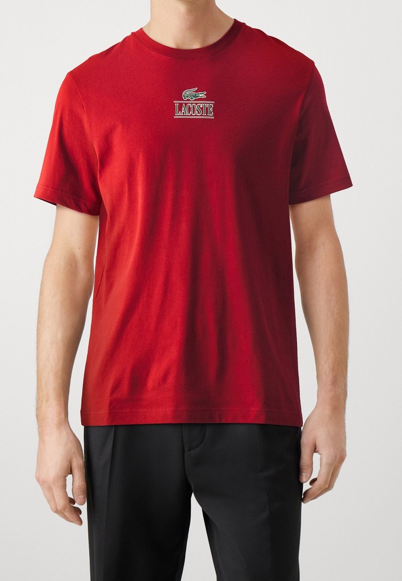 Homme portant un t-shirt rouge à manches courtes avec un petit logo crocodile vert et le texte « LACOSTE » sur la poitrine, associé à un pantalon noir.