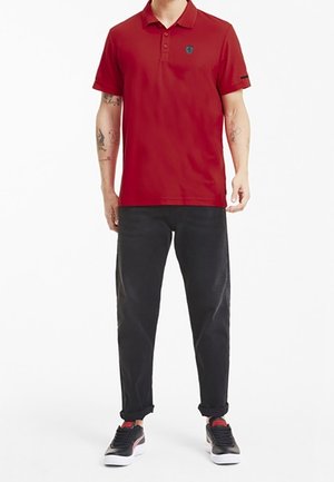 Uomo che indossa una polo rossa a maniche corte, pantaloni neri risvoltati e sneakers nere con suole bianche, in piedi contro uno sfondo bianco.